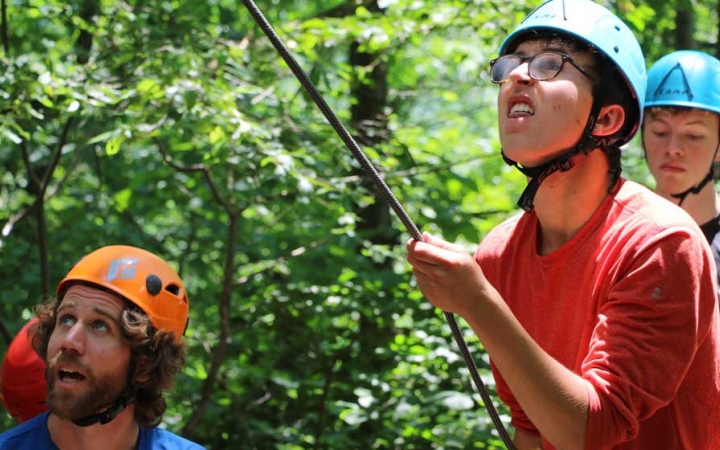 Three people wearing helmets look up at an apparent rock climber while standing in a wooded area. One of them appears to be belaying the climber. 